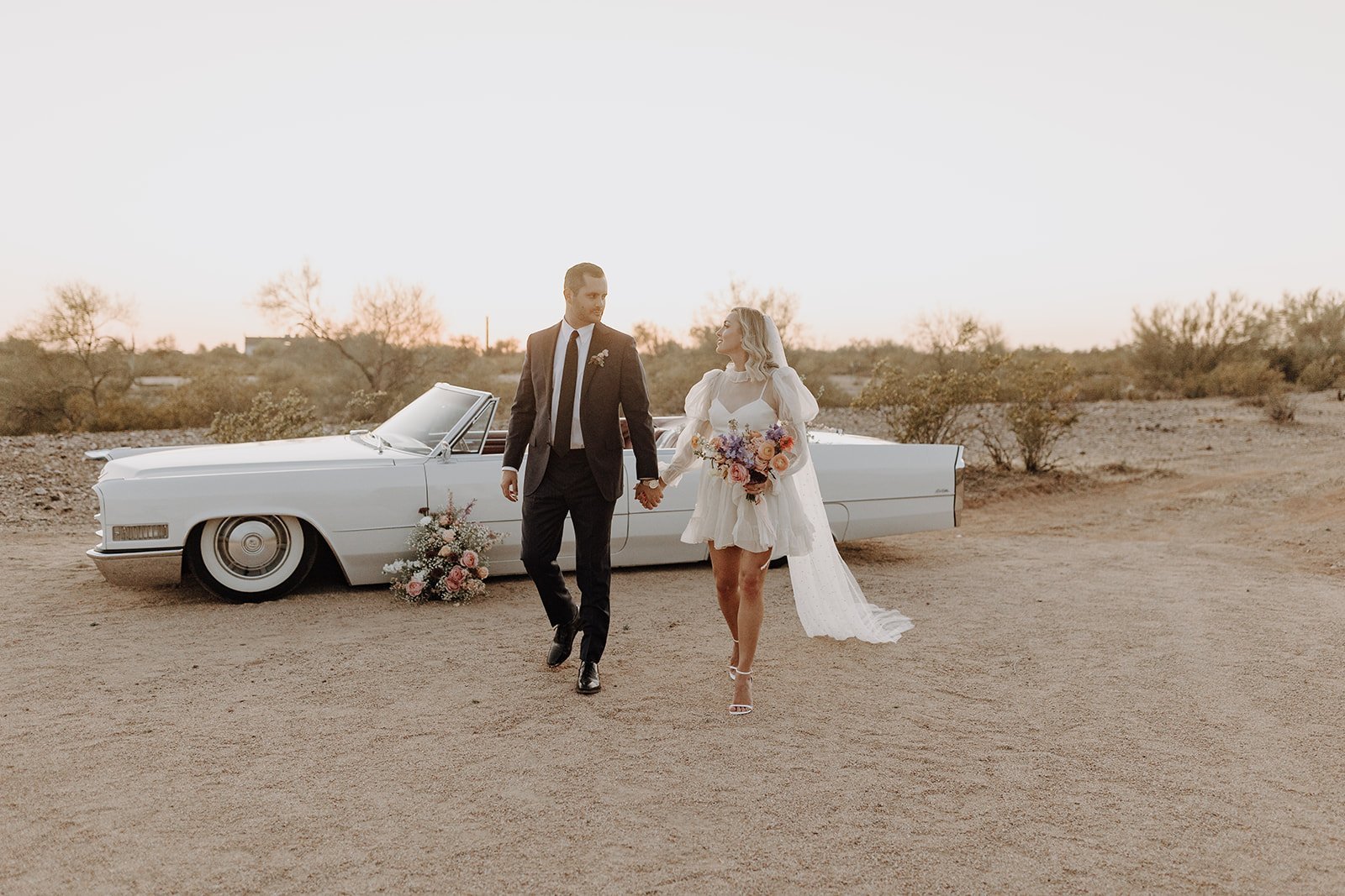 Bride and groom walking hand in hand next to vintage white convertible for wedding styled shoot