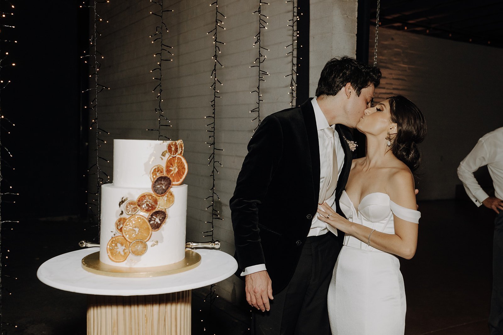 Bride and groom kissing next to their white two-tier wedding cake decorated with dried oranges