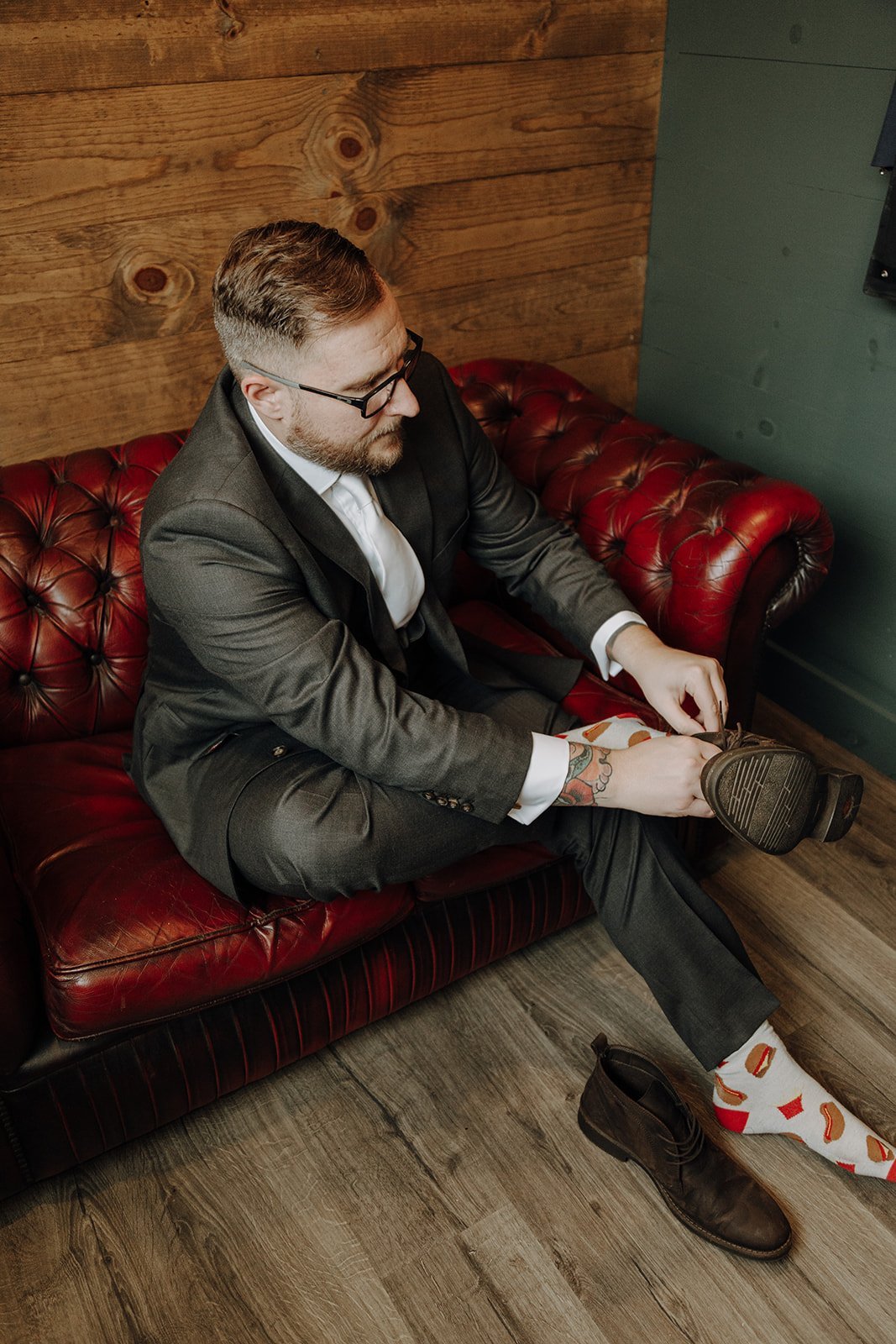 Groom wearing black suit and hamburger socks, sitting on a brown leather couch and tying his shoes