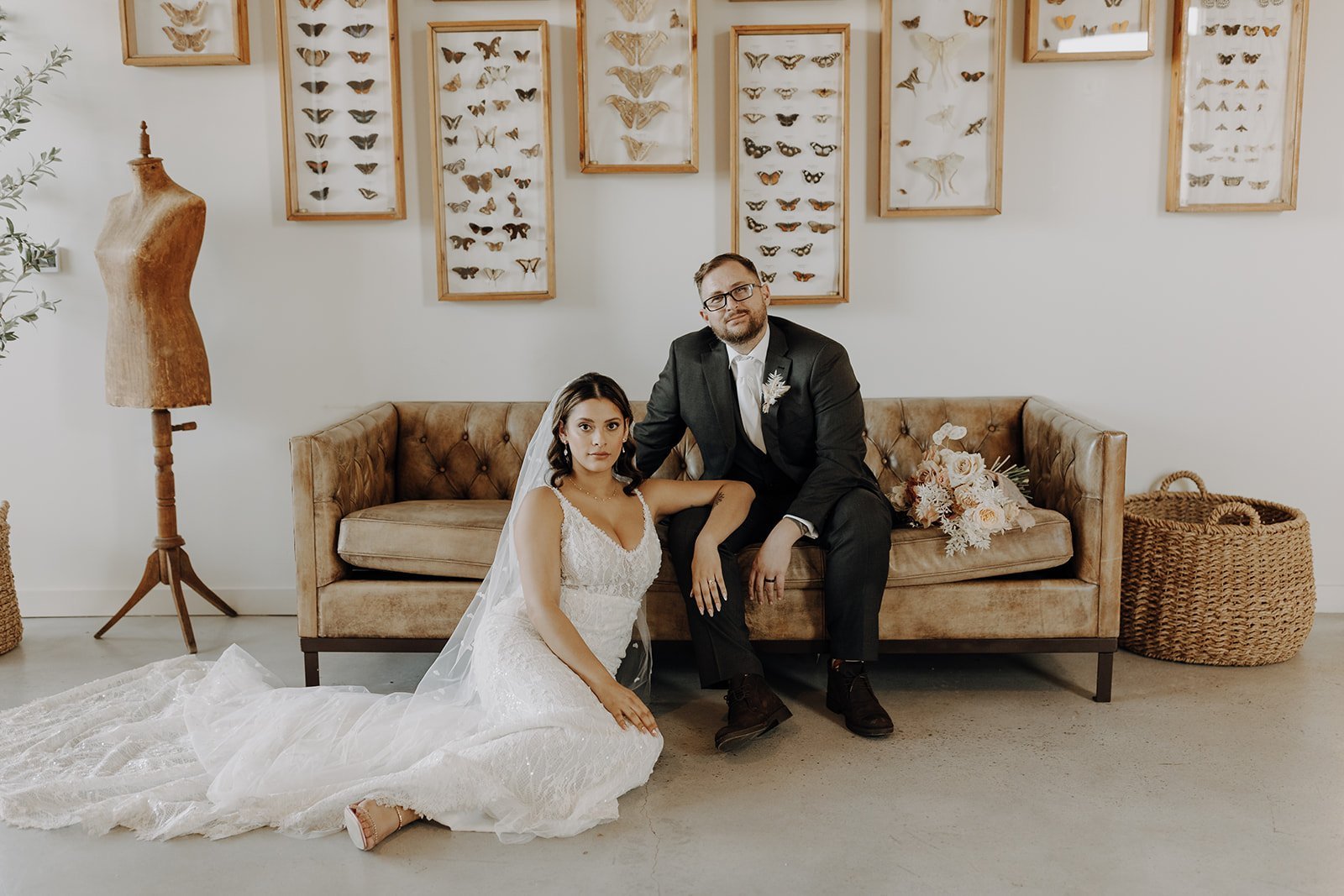 Groom sitting on brown leather couch while bride sits on the floor, posing for couple photos