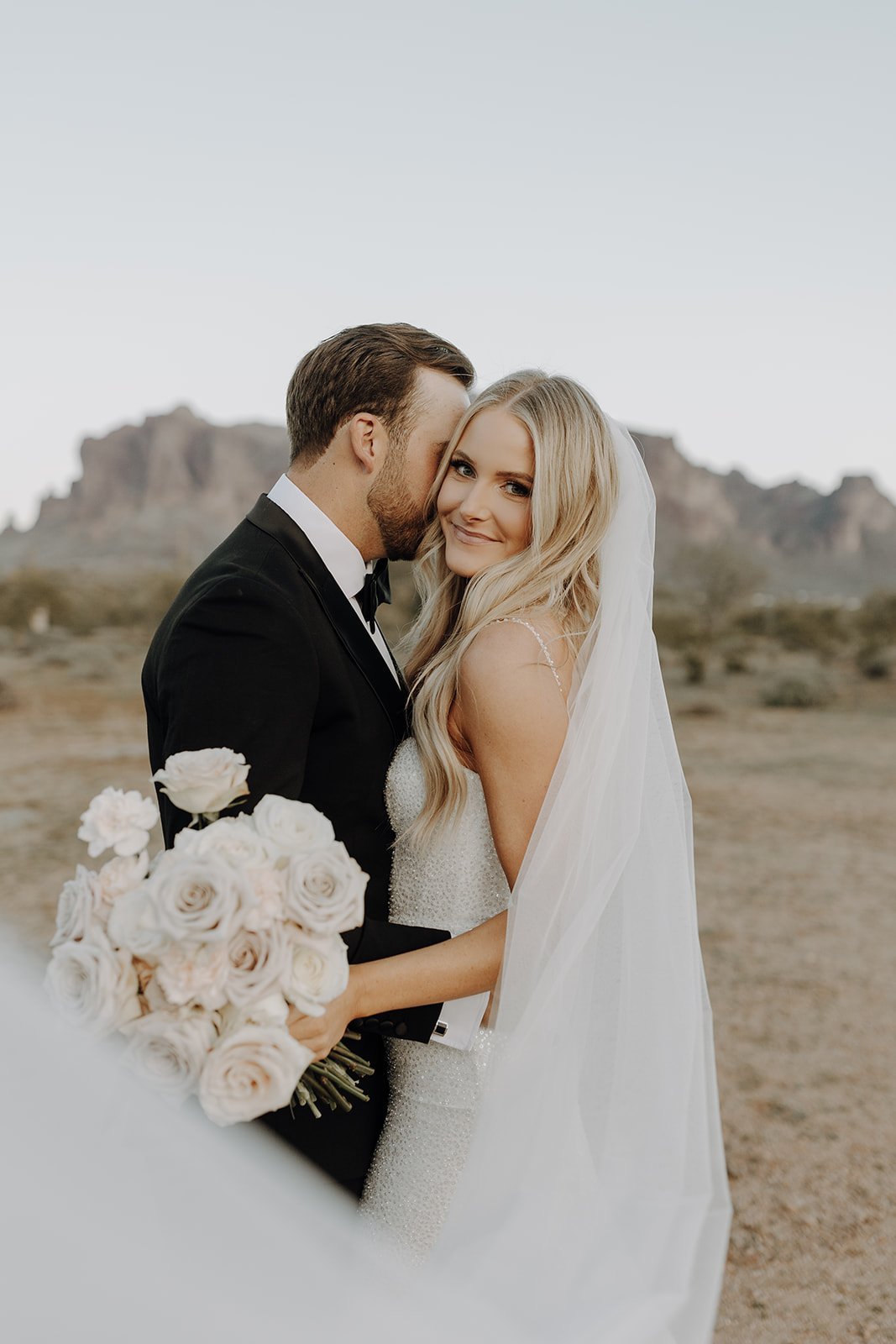Groom kissing bride in the desert while she smiles at the camera and holds a white bouquet