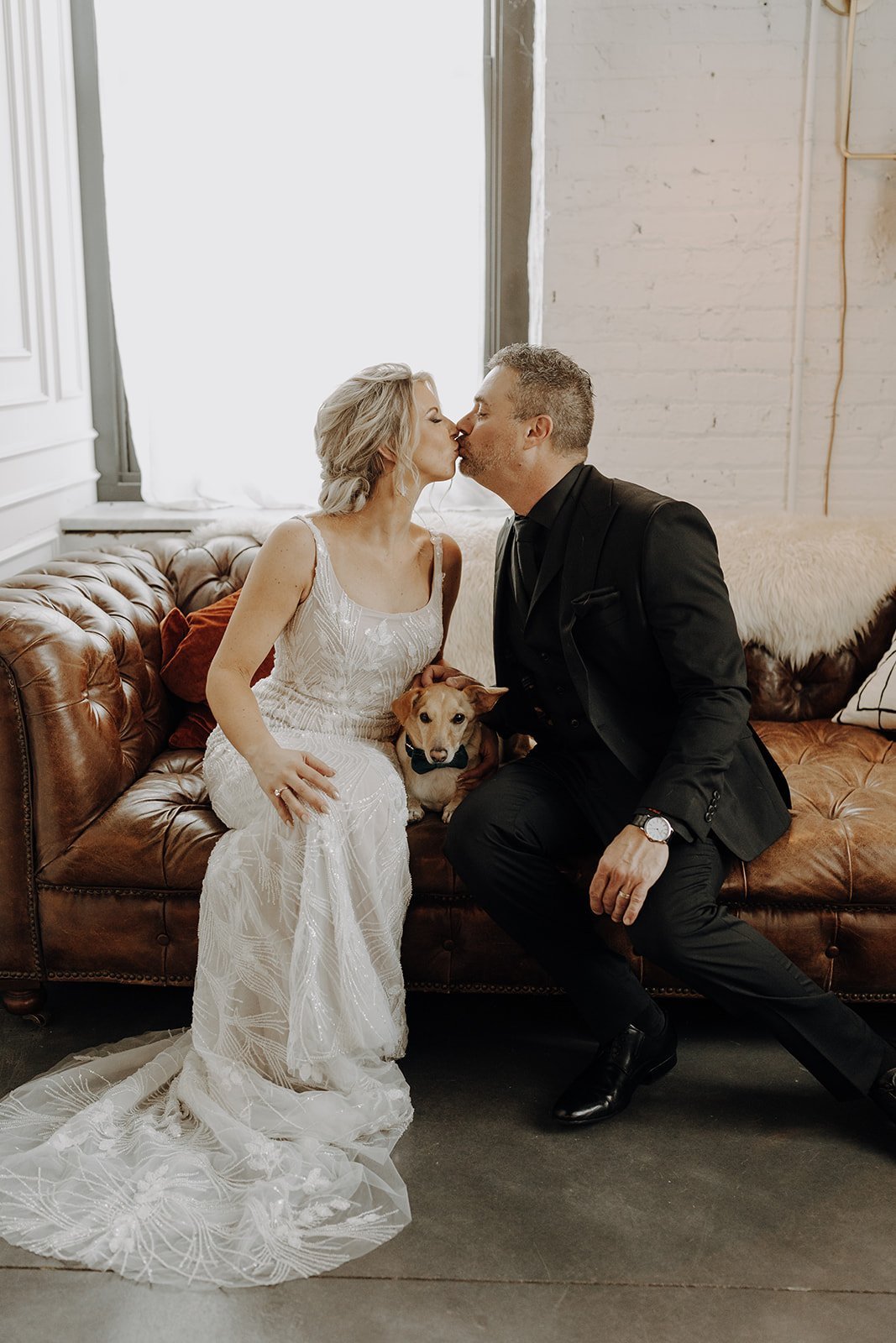 Bride and groom sitting and kissing on brown leather couch at non-traditional wedding in Illinois