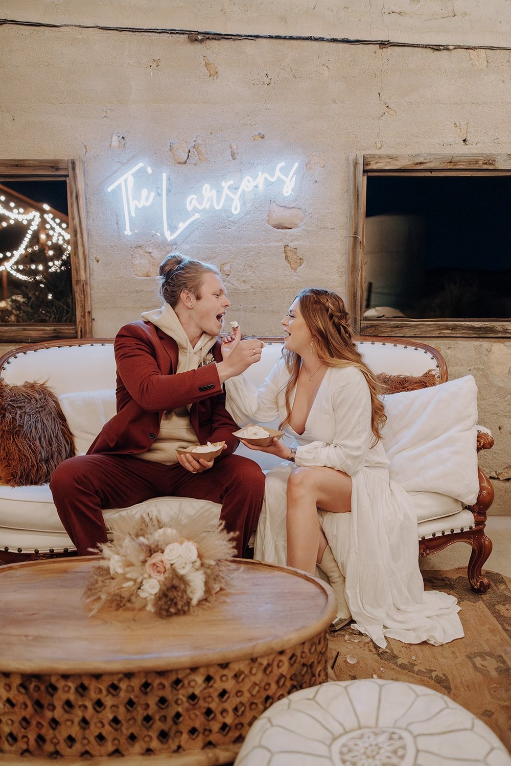 Bride and groom sit on a white couch and feed each other cake at their Joshua Tree wedding