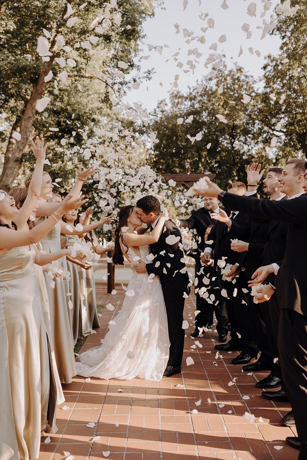 Bride and groom kiss while wedding party throws white petals