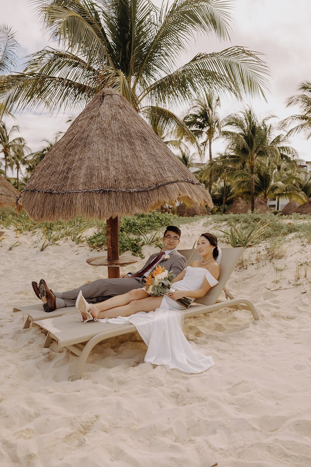 Bride and groom recline on lounge chairs on the beach before their wedding ceremony