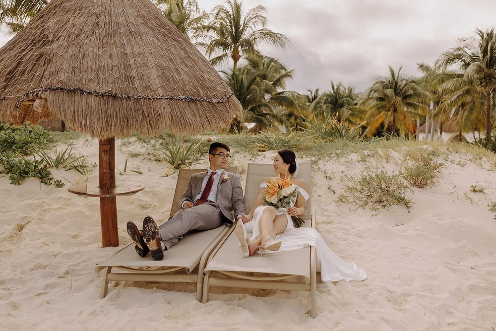 Bride and groom recline on lounge chairs on the beach before their wedding ceremony