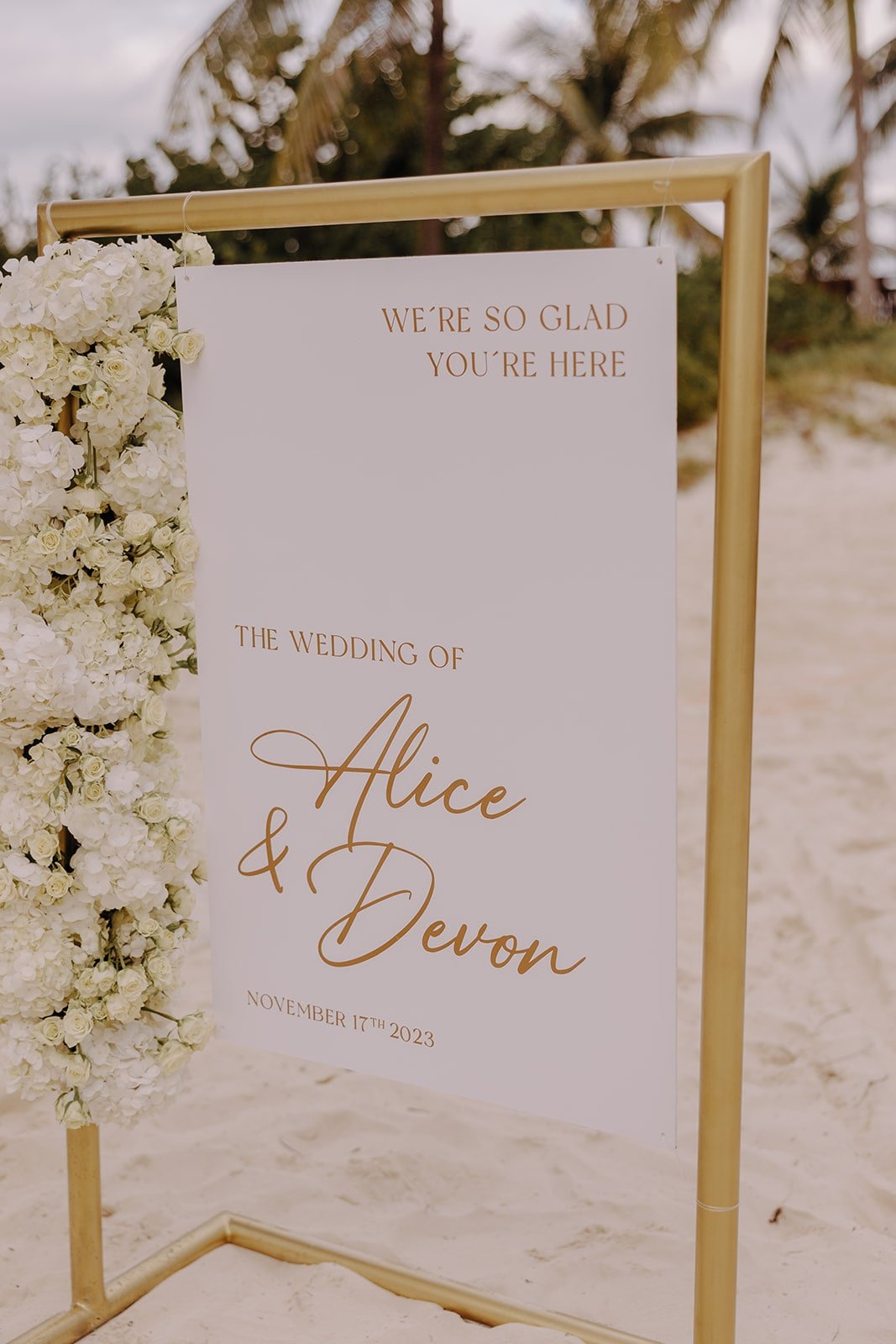 White wedding welcome sign hanging from a gold frame on the beach