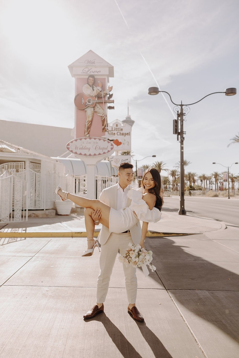 Groom picks up bride for a photo in front of the Little White Chapel sign during their Las Vegas elopement