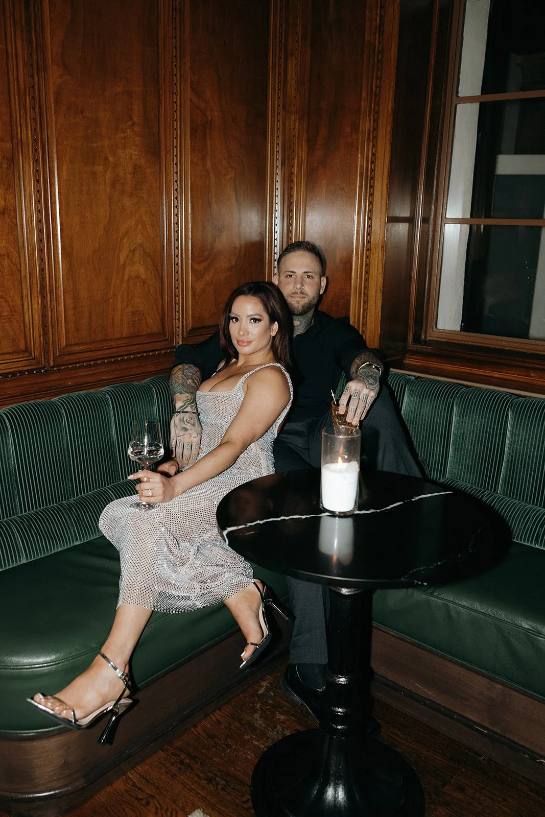 A woman in a sparkly dress and a man in dark clothing sit together on a green bench in a wood-paneled room for their luxury engagement photos