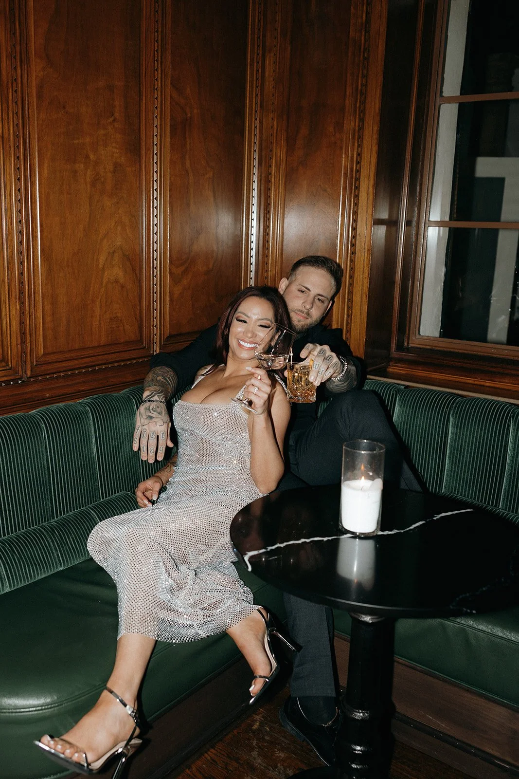 A woman in a sparkly dress and a man in dark clothing sit together on a green bench in a wood-paneled room for their luxury engagement photos