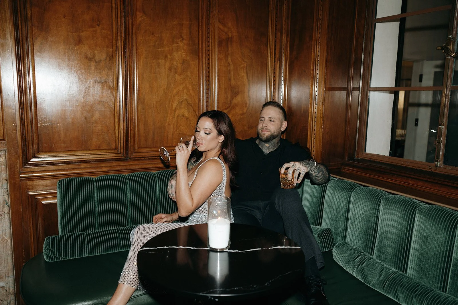A woman in a sparkly dress and a man in dark clothing sit together on a green bench in a wood-paneled room for their luxury engagement photos