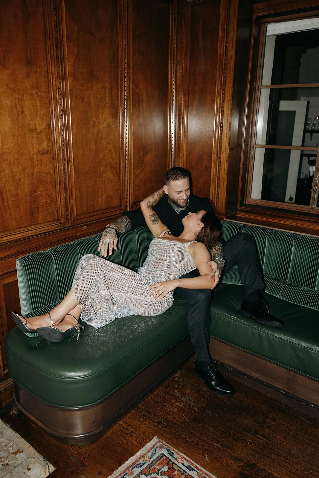 A woman in a sparkly dress and a man in dark clothing sit together on a green bench in a wood-paneled room for their luxury engagement photos