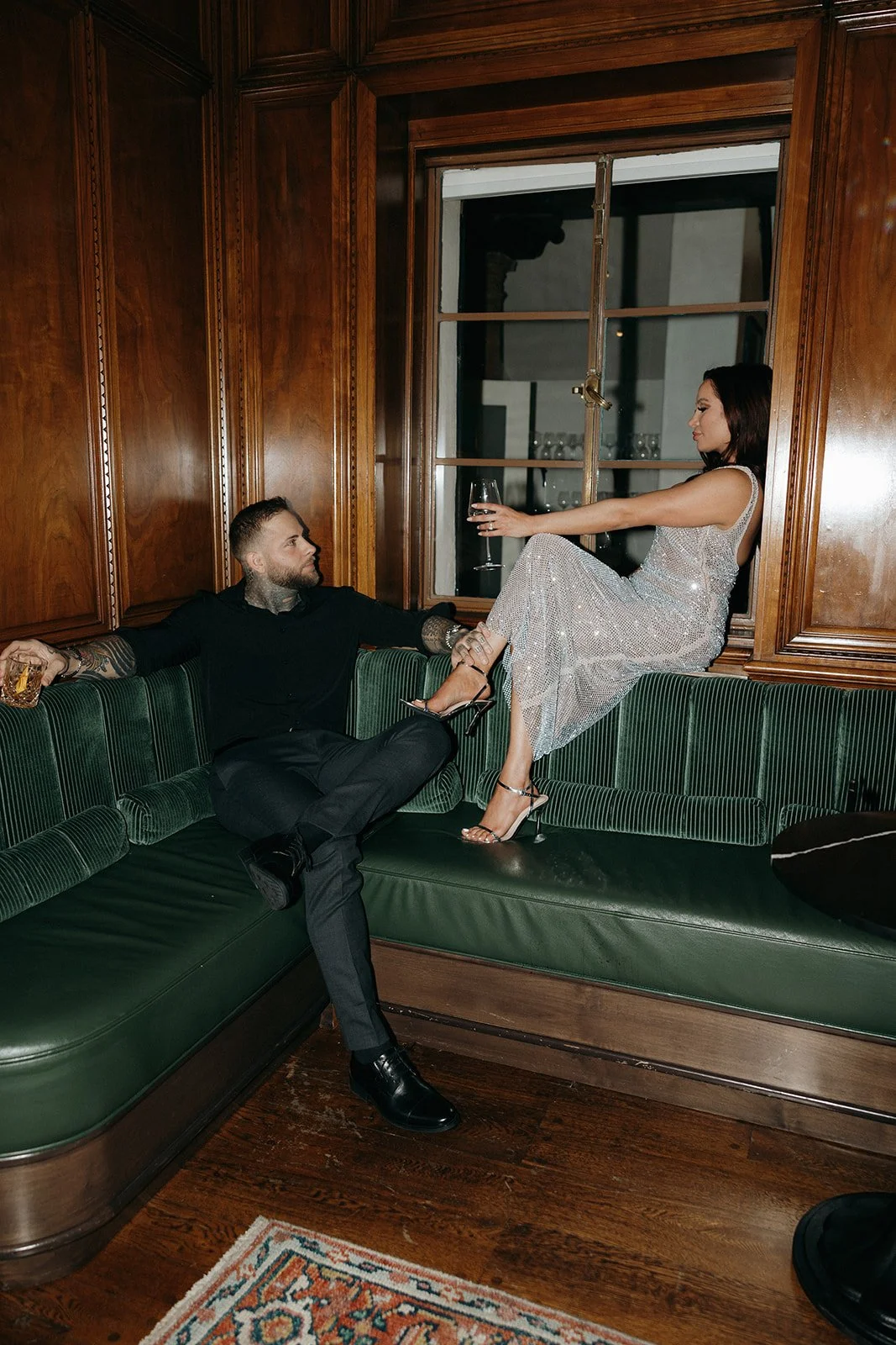 A woman in a sparkly dress and a man in dark clothing sit together on a green bench in a wood-paneled room for their luxury engagement photos