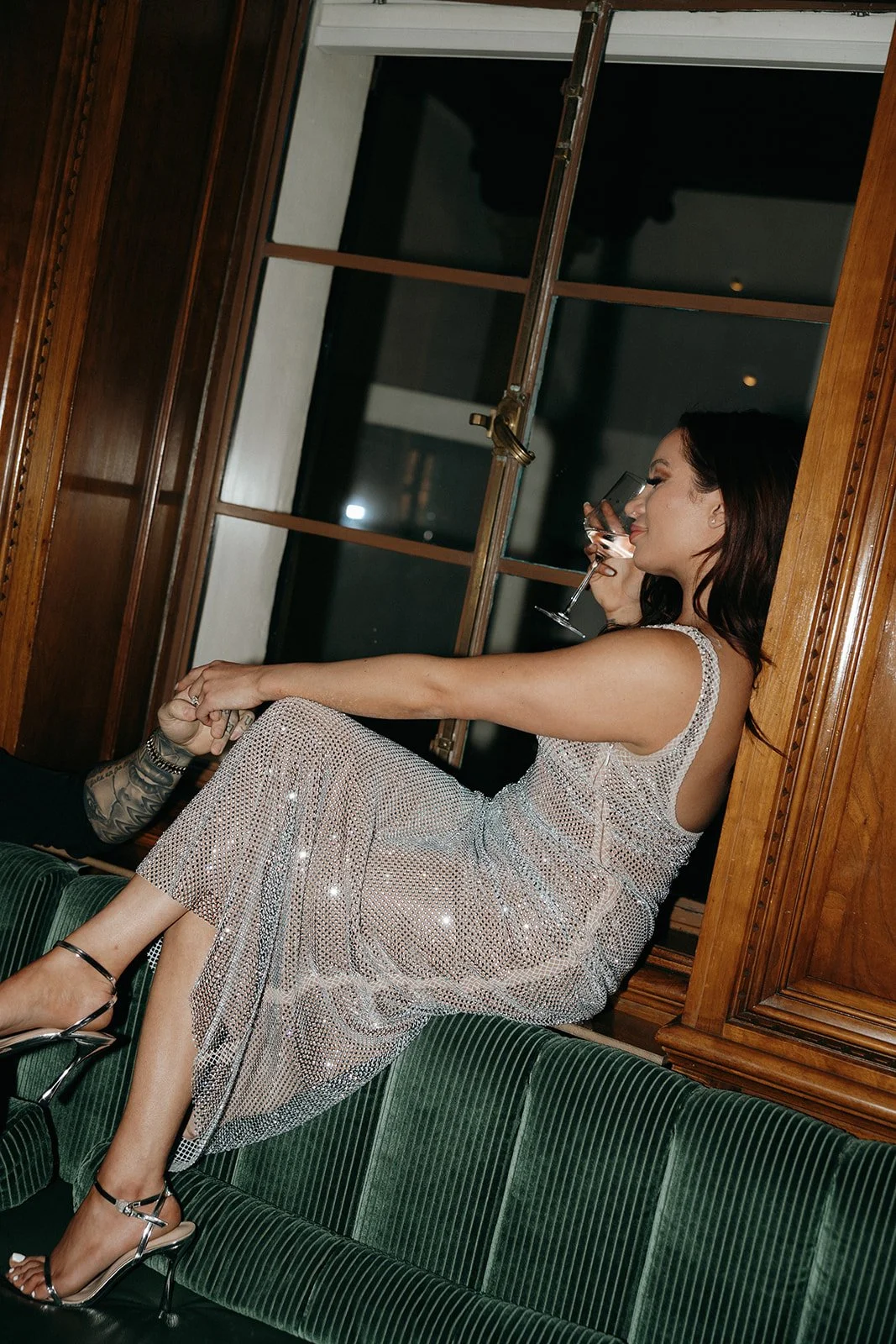A woman in a sparkly dress and a man in dark clothing sit together on a green bench in a wood-paneled room for their luxury engagement photos