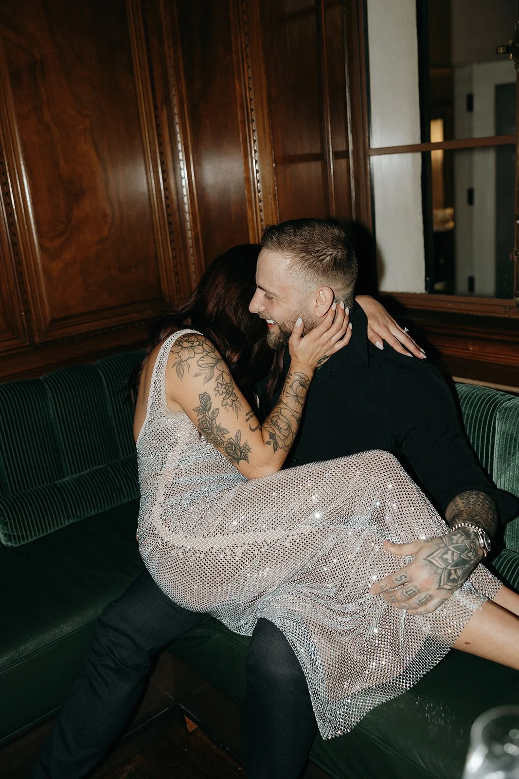 A woman in a sparkly dress and a man in dark clothing sit together on a green bench in a wood-paneled room for their luxury engagement photos