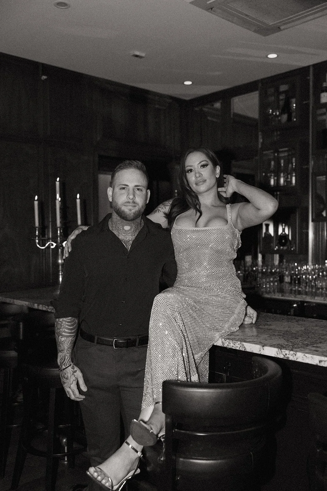 A woman in a sparkly dress and a man in dark clothing sit together on a green bench in a wood-paneled room for their luxury engagement photos