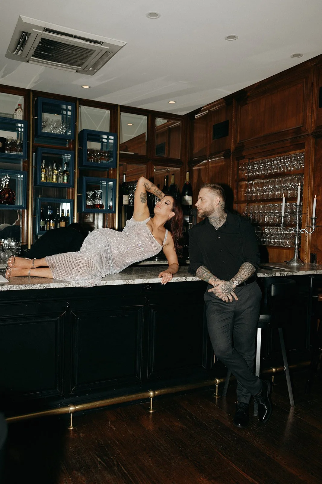 A woman in a sparkly dress and a man in dark clothing sit together on a green bench in a wood-paneled room for their luxury engagement photos