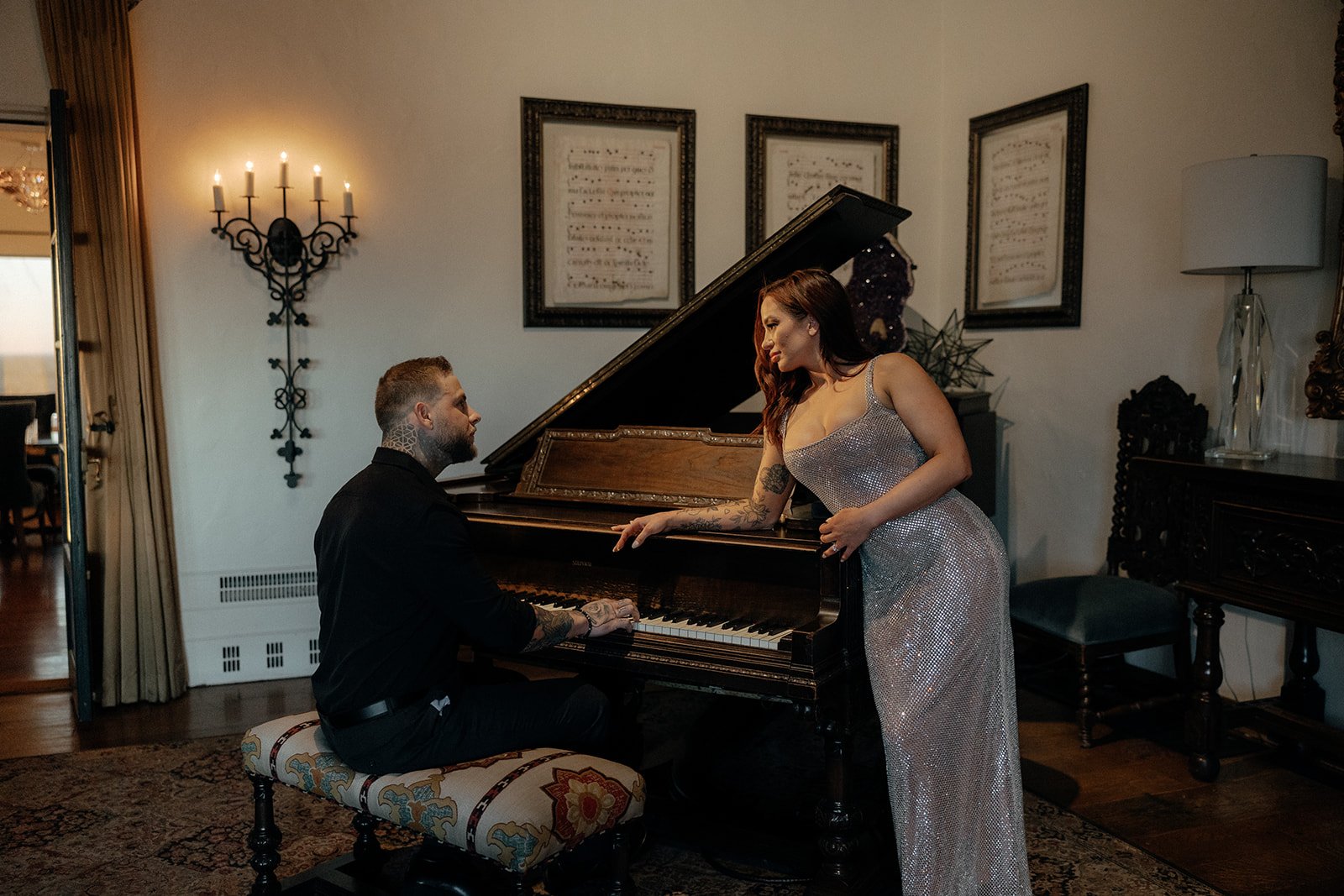 A man plays a grand piano while a woman in a formal dress leans on it, both in an elegantly decorated room for their luxury engagement photos