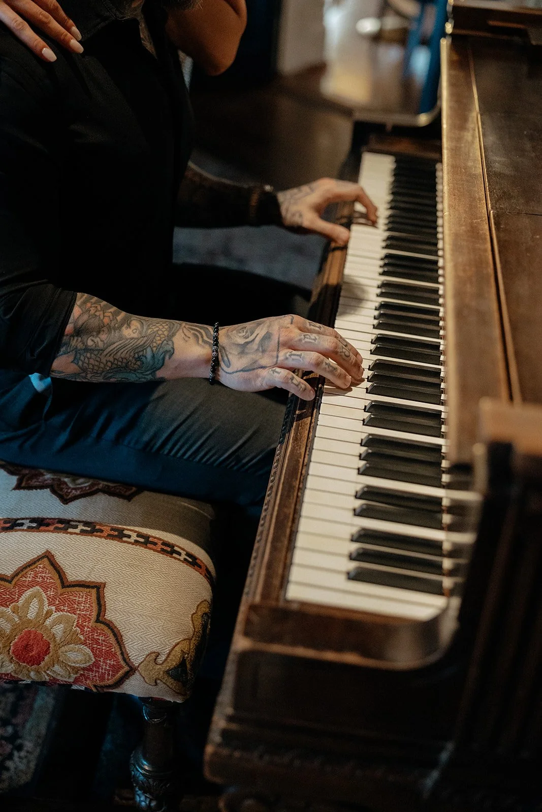 A person with tattooed arms plays a piano, sitting on a patterned cushioned bench.
