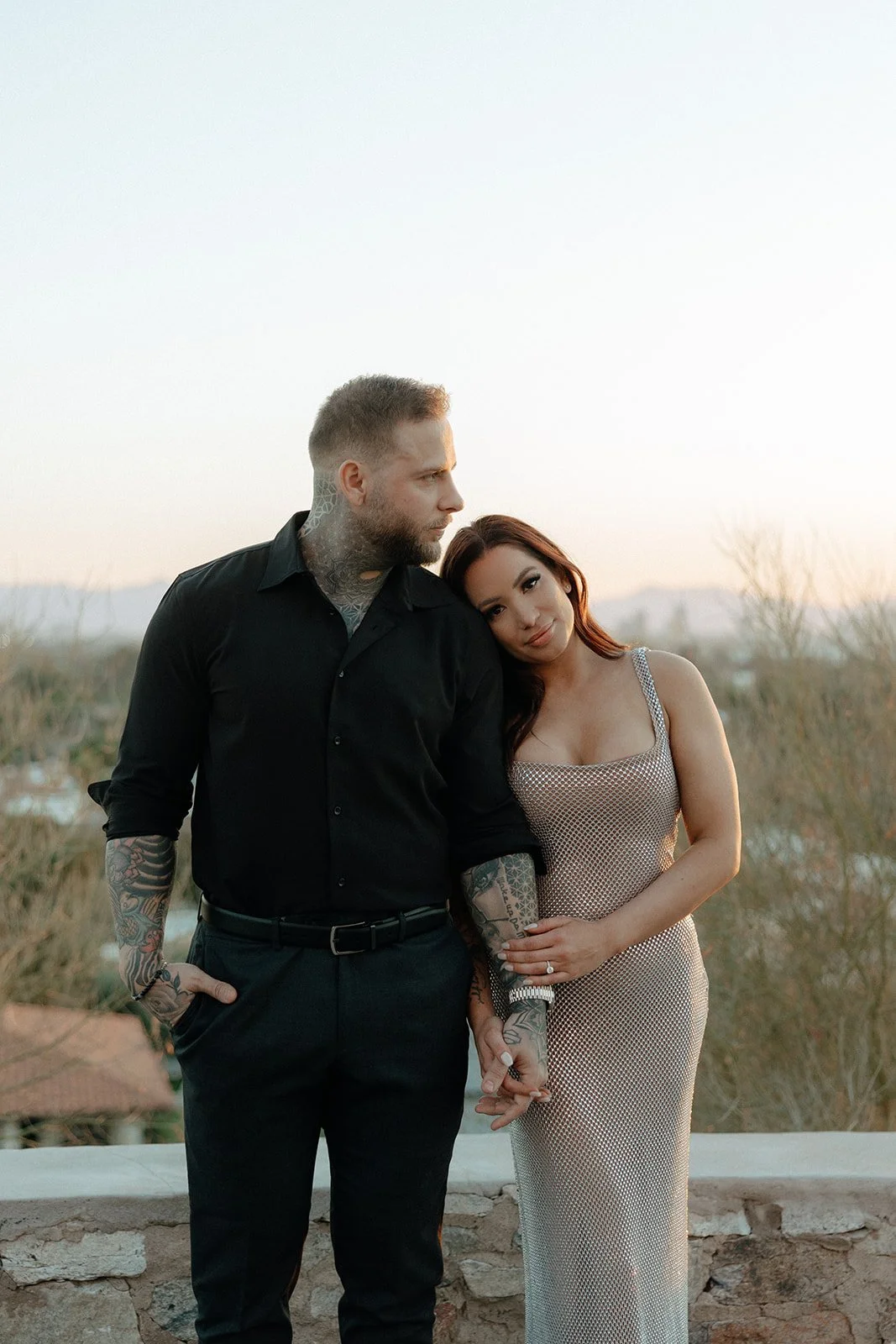 A man and woman stand outdoors holding hands; the woman leans her head on the man's shoulder for their luxury engagement photos