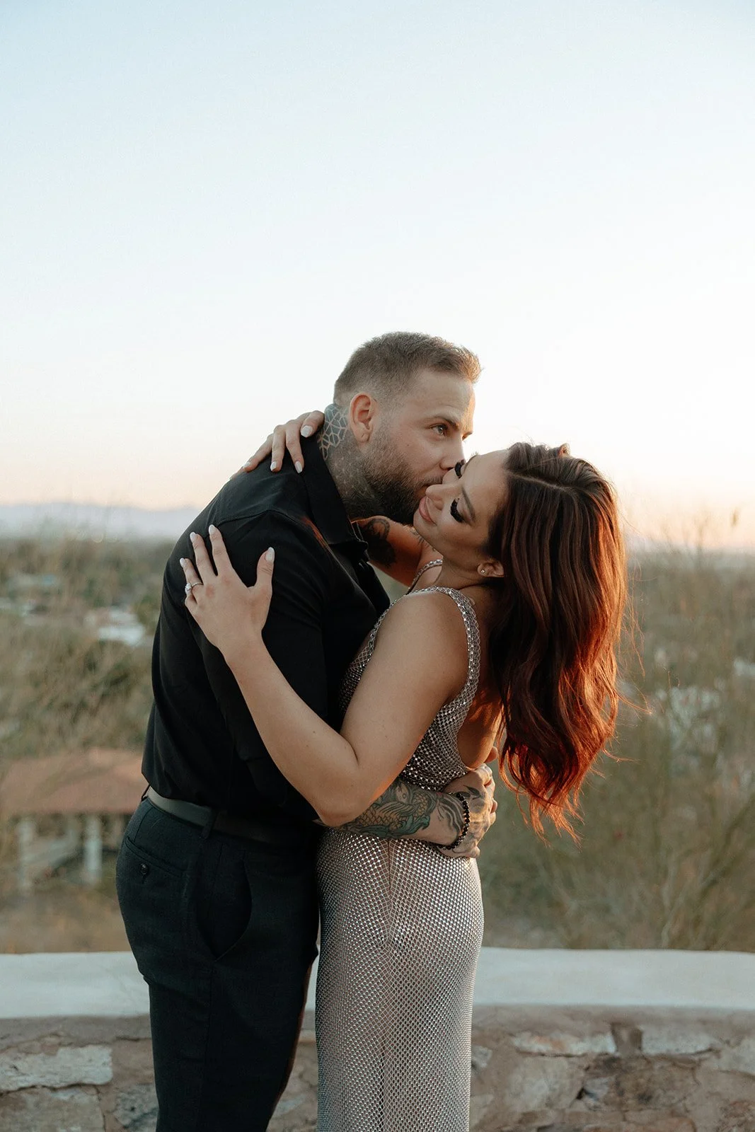 A man and woman stand outdoors holding hands; the woman leans her head on the man's shoulder for their luxury engagement photos