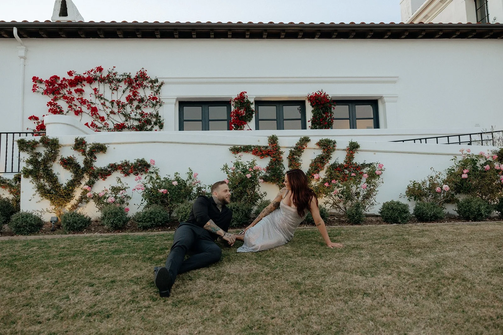 A couple sits on the grass in front of a white building with flower-covered walls and windows.