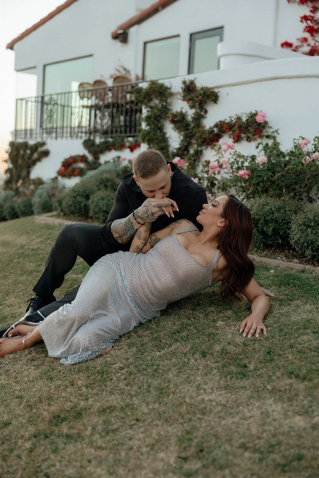 A couple sits on the grass in front of a white building with flower-covered walls and windows for their luxury engagement photos