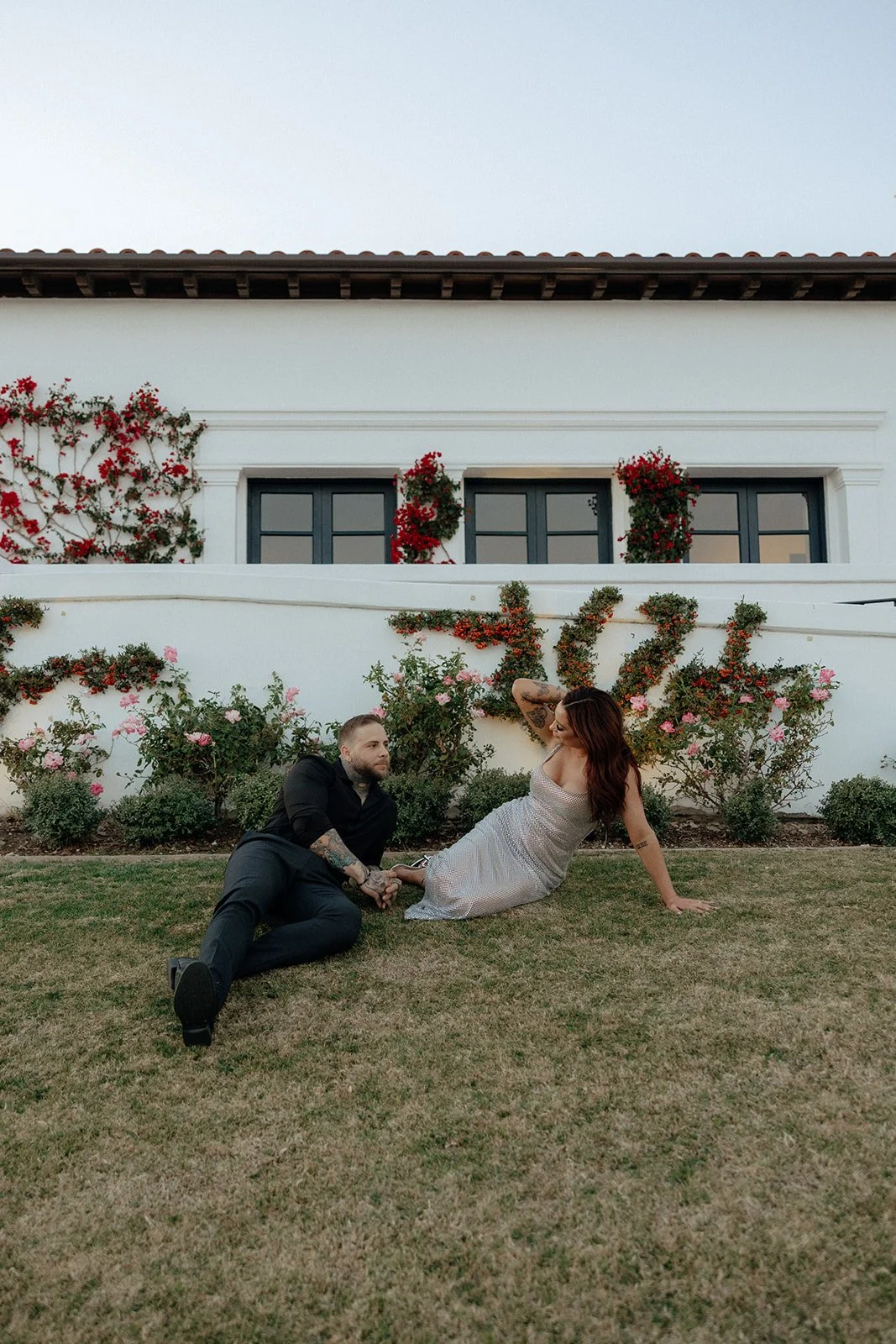 A couple sits on the grass in front of a white building with flower-covered walls and windows for their luxury engagement photos