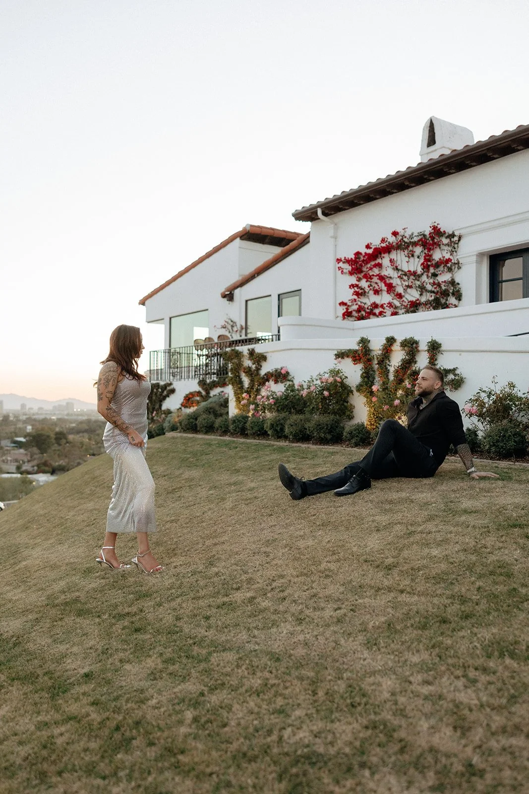 A couple sits on the grass in front of a white building with flower-covered walls and windows for their luxury engagement photos