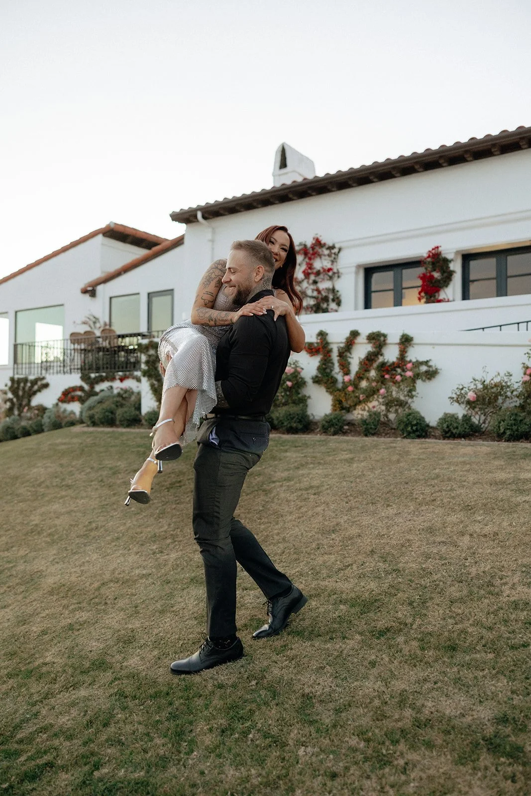 A couple sits on the grass in front of a white building with flower-covered walls and windows for their luxury engagement photos