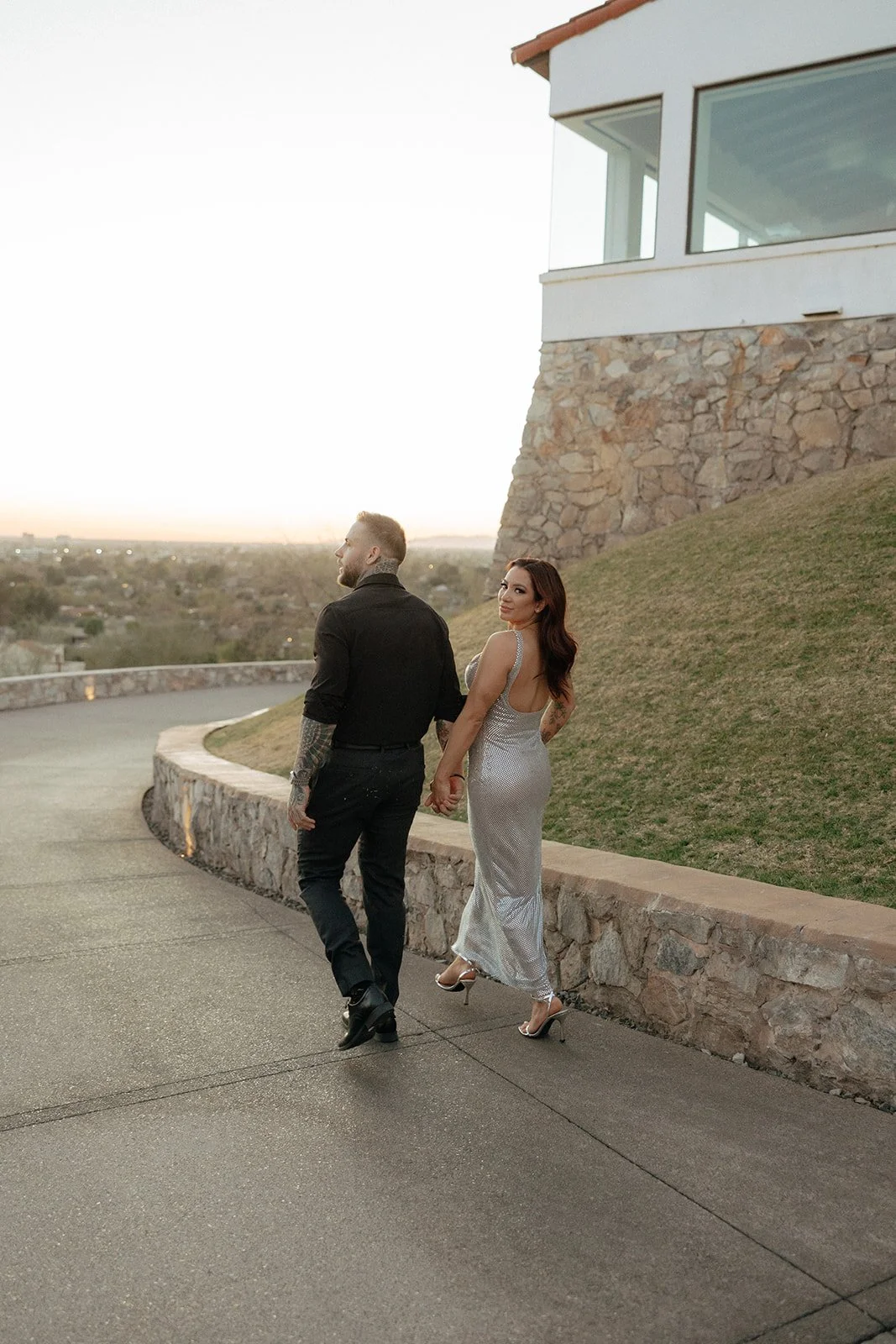 A man in black clothing and a woman in a silver dress walk hand in hand on a curved path near a stone wall and grassy hill at sunset.