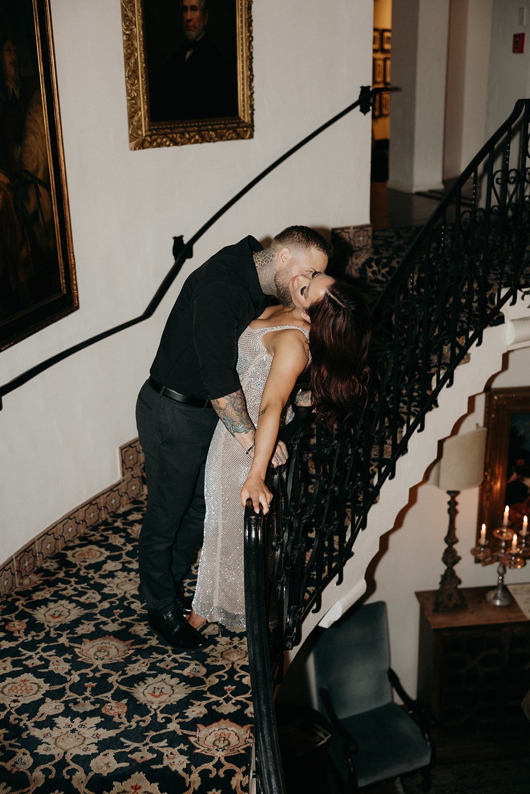 A man in black clothing and a woman in a sparkly dress stand on a carpeted, curved staircase with ornate railings and framed portraits on the wall.