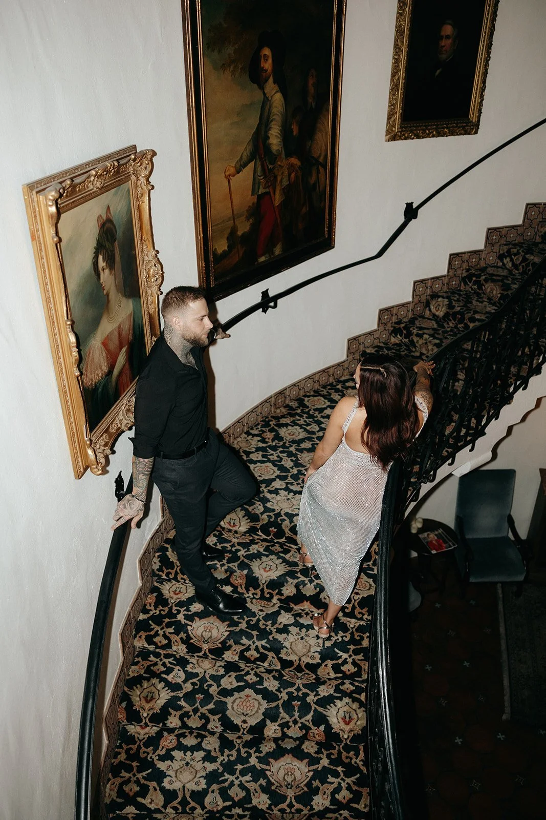 A man in black clothing and a woman in a sparkly dress stand on a carpeted, curved staircase with ornate railings and framed portraits on the wall.