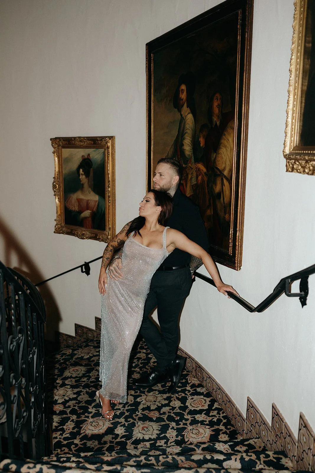A man in black clothing and a woman in a sparkly dress stand on a carpeted, curved staircase with ornate railings and framed portraits on the wall.