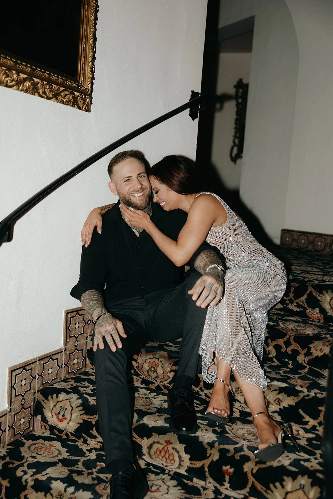 A woman in a sparkling dress sits on a staircase next to a man in black attire, smiling and leaning affectionately against him.