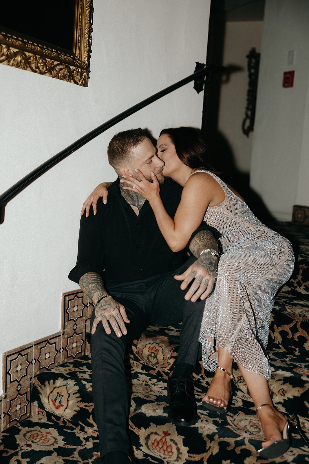 A woman in a sparkling dress sits on a staircase next to a man in black attire, smiling and leaning affectionately against him.