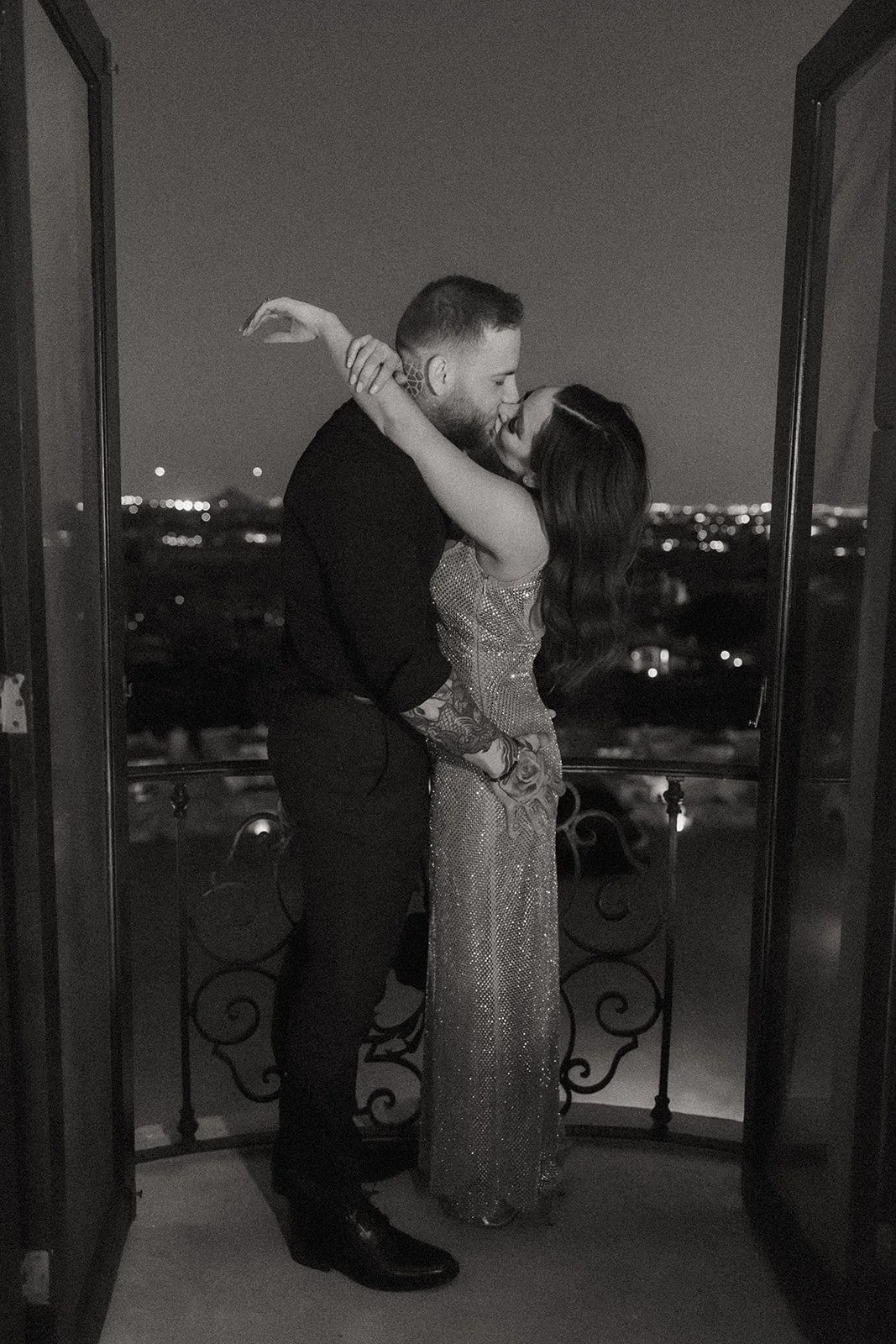 A couple stands on a balcony at night, embracing and kissing, with city lights visible in the background.