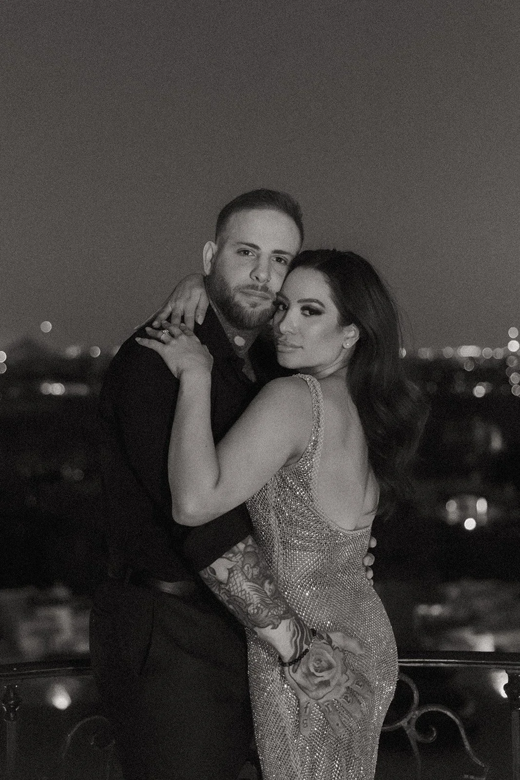 A couple stands on a balcony at night, embracing and kissing, with city lights visible in the background.