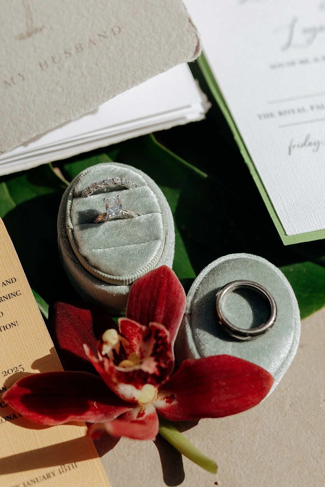Two arch-shaped cards, one pink and one beige, displaying wedding event details, placed on a surface with flowers and green leaves nearby for a destination tropical wedding