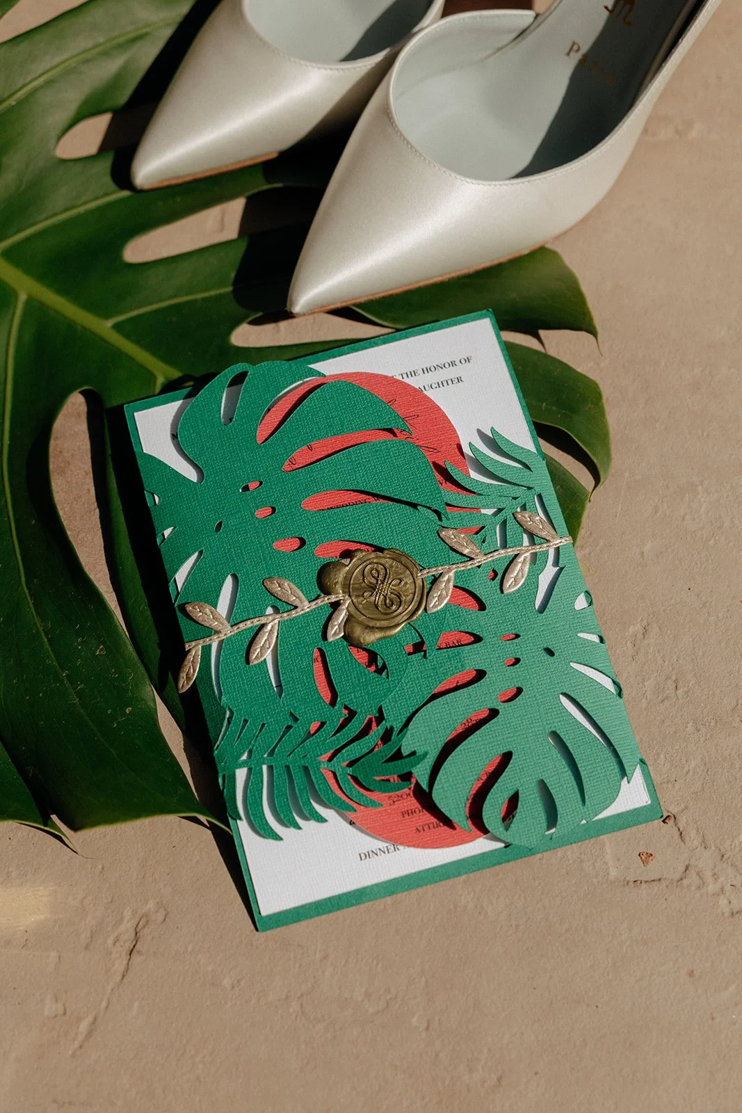 A decorative green tropical-themed wedding invitation with a wax seal, placed on a monstera leaf, next to a pair of white pointed-toe shoes.