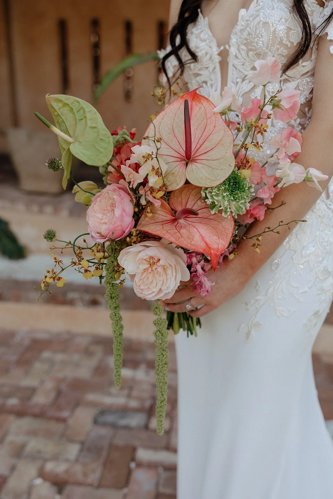 A bride in a white, off-shoulder wedding dress holds a colorful bouquet and smiles while standing outdoors near stone columns and greenery for destination tropical wedding 