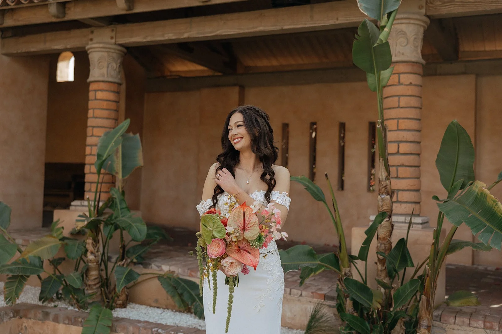 A bride in a white, off-shoulder wedding dress holds a colorful bouquet and smiles while standing outdoors near stone columns and greenery for destination tropical wedding 