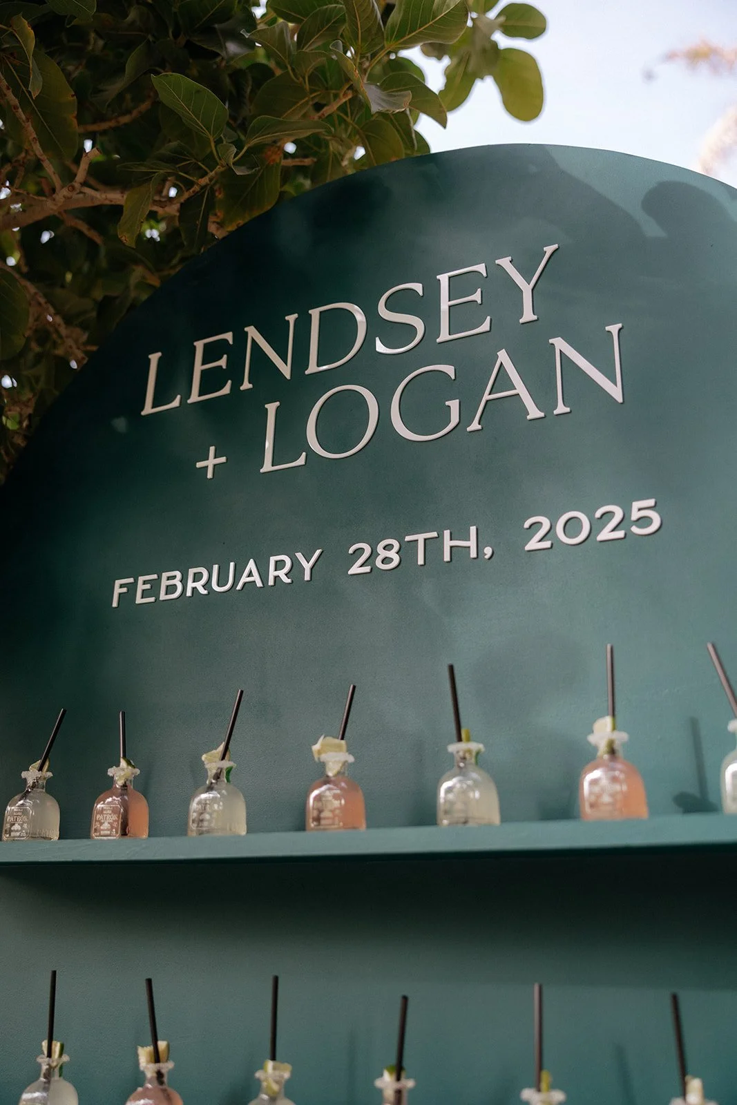 A green wedding display board with “Lendsey + Logan, February 26th, 2025” holds rows of small bottles, decorated with tropical flowers at the base.