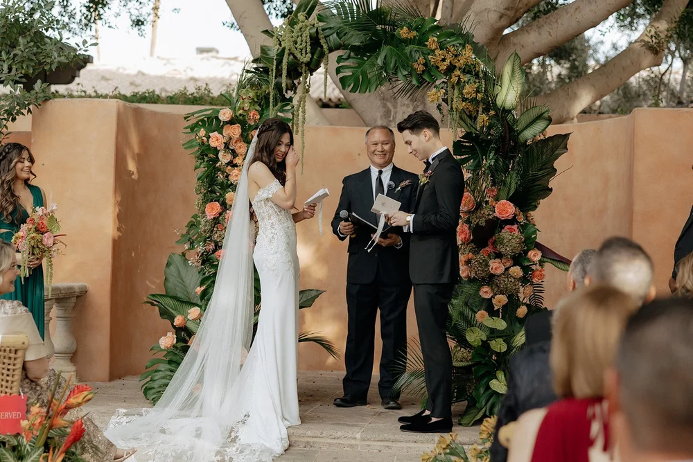 A bride and groom stand under a floral arch exchanging vows, with an officiant between them, outdoors for a destination tropical wedding 