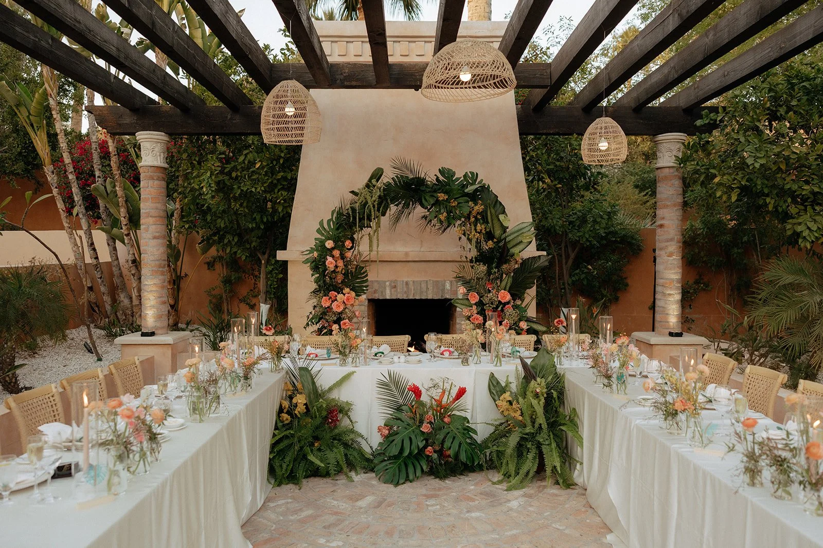 Outdoor dining table set for an event with white tablecloths, floral centerpieces, and greenery, arranged under a pergola in front of a decorated fireplace.