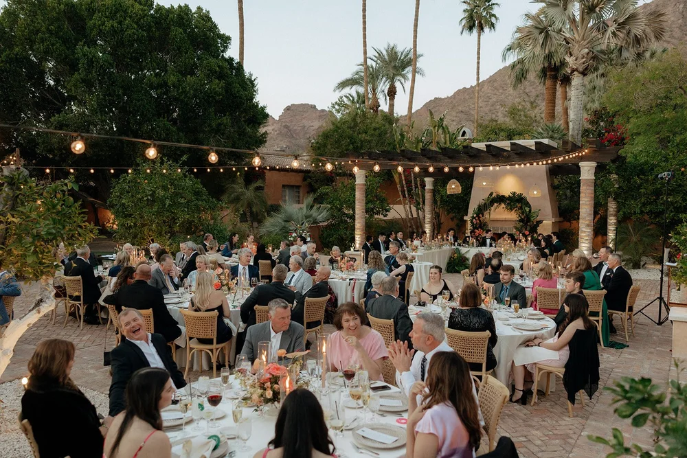 Large group of people seated at round tables outdoors for a formal event, with string lights overhead and palm trees in the background.