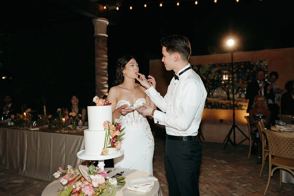 A bride and groom cut their wedding cake together at an outdoor evening reception.