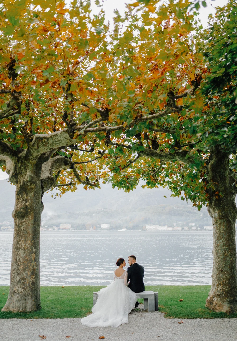 A bride and groom walk hand in hand across a manicured lawn in a scenic outdoor garden with mountains and water in the background for a destination wedding in lake como