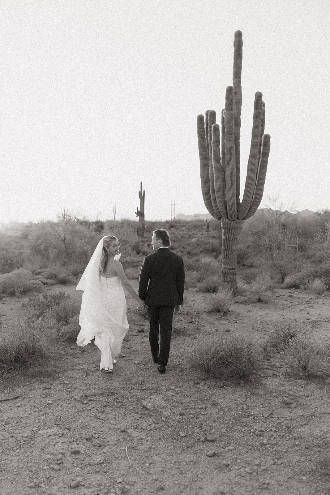 A bride and groom embrace in wedding attire in a desert landscape, with a tall cactus in the background