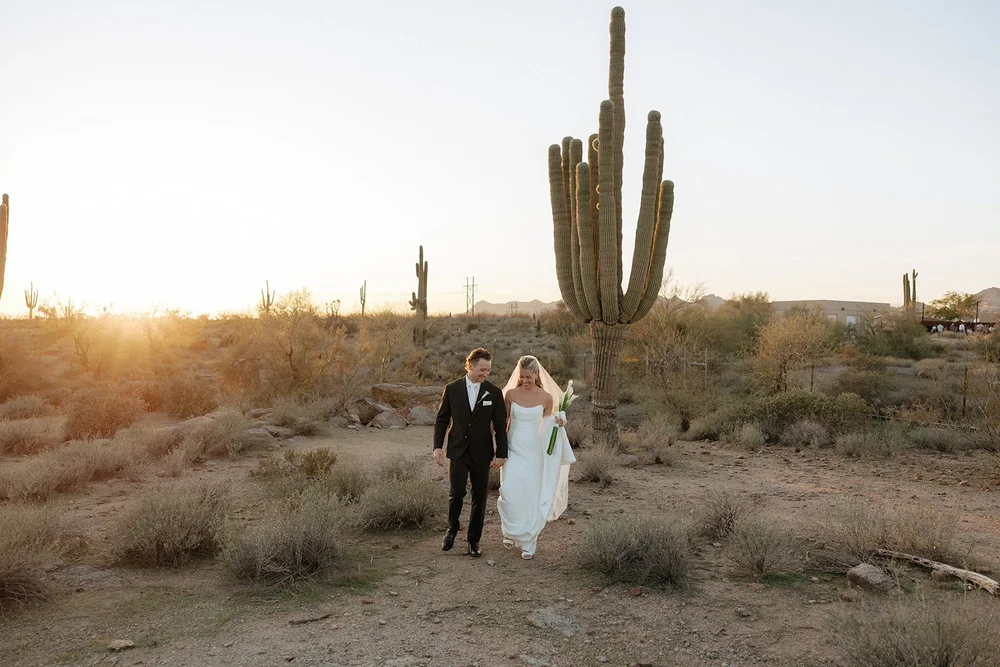 A bride and groom embrace in wedding attire in a desert landscape, with a tall cactus in the background